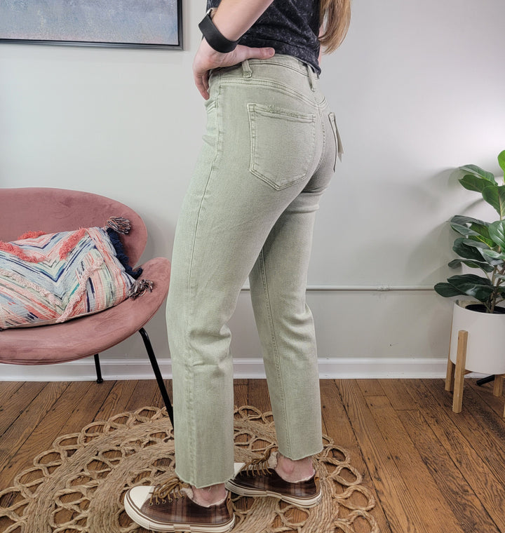 Wearing Lovervet’s Ariel Olive Straight Jeans, a person stands on a woven rug in brown sneakers. The cropped straight-leg denim is visible alongside a blush pink chair, vibrant pillow, and potted plant. Only the lower half shows.