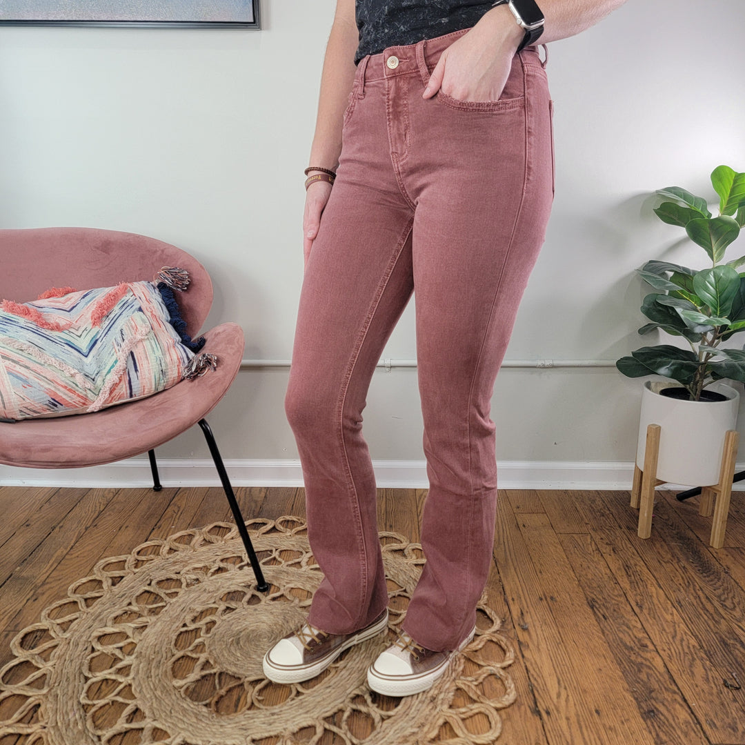 A person stands indoors on a circular woven rug, wearing Lovervet's Angela Wine Bootcut Jeans with comfort stretch denim, a black top, sneakers, and a smartwatch. A pink chair, plant, and wooden floor complete the cozy background.