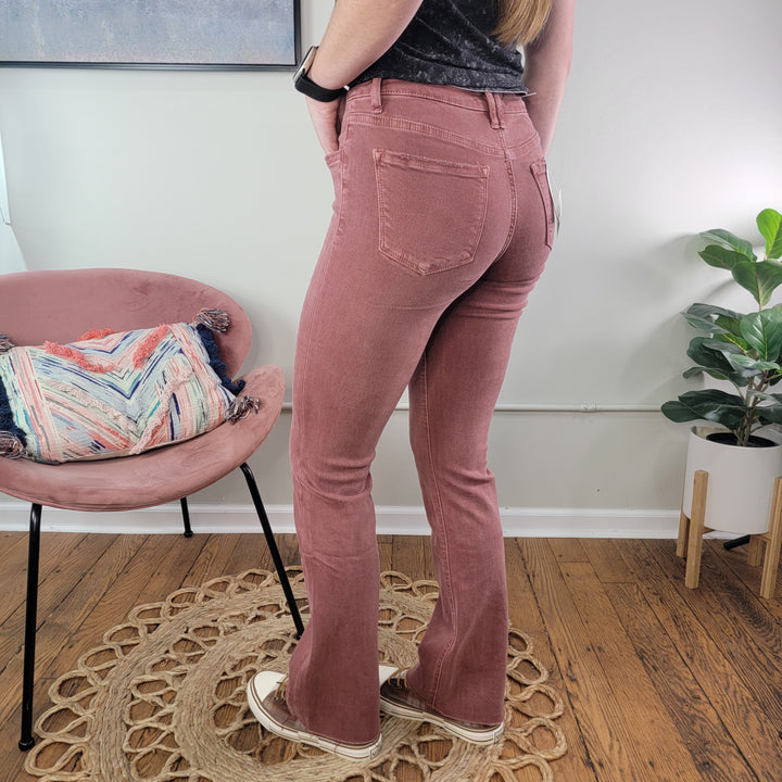 A person stands indoors on a woven rug beside a pink chair, wearing Lovervet’s Angela Wine Bootcut Jeans in comfort stretch denim and cream sneakers, with a colorful pillow and potted plant nearby.