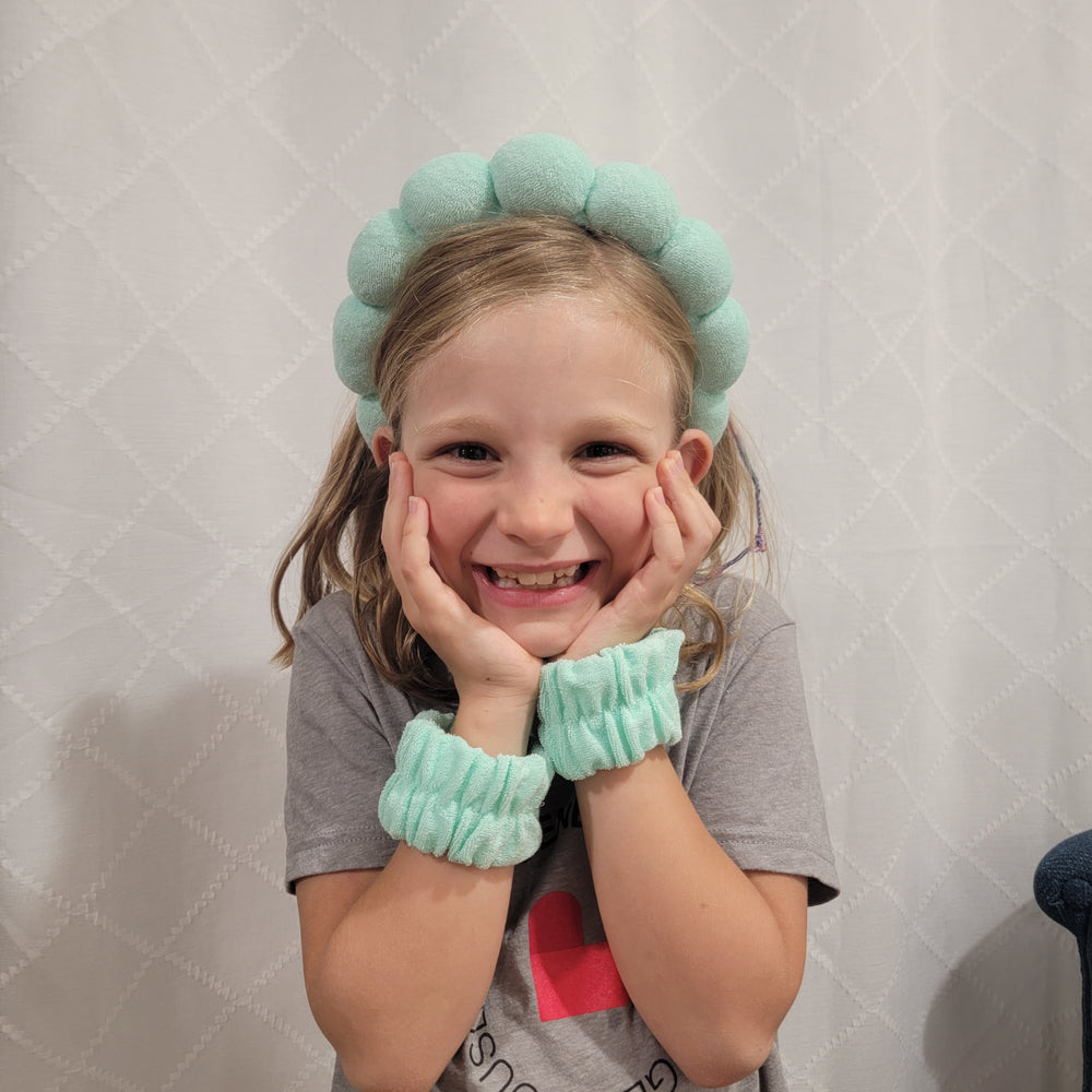 A young blonde girl smiles, resting her face in her hands. She wears the DM Merchandising "Washed Up Like This" Dryband Set—gray shirt, padded headband, and matching blue terry cloth wristbands—against a white textured background.