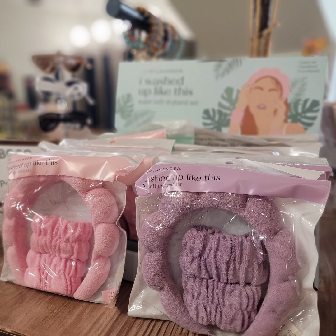 The "Washed Up Like This" Dryband Set by DM Merchandising features two pink and purple terry cloth headbands in clear packaging on a wooden surface, with a blurred sign and other accessories in the background.