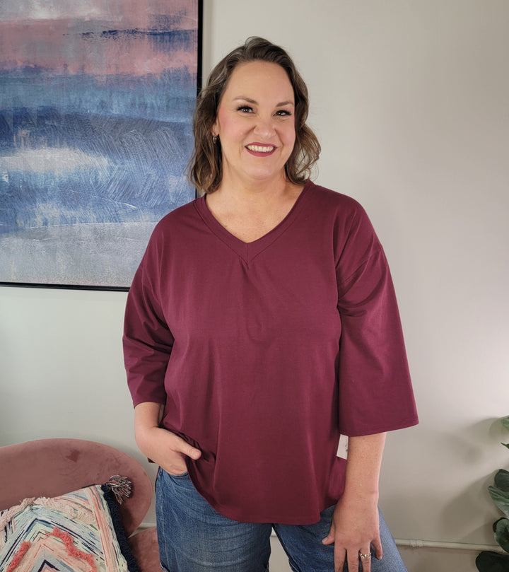 A woman with wavy brown hair smiles indoors, wearing the Sew In Love Esme V-neck top in burgundy with wide 3/4 sleeves and blue jeans. Behind her are abstract blue and pink wall art and a pink chair draped with a patterned blanket.