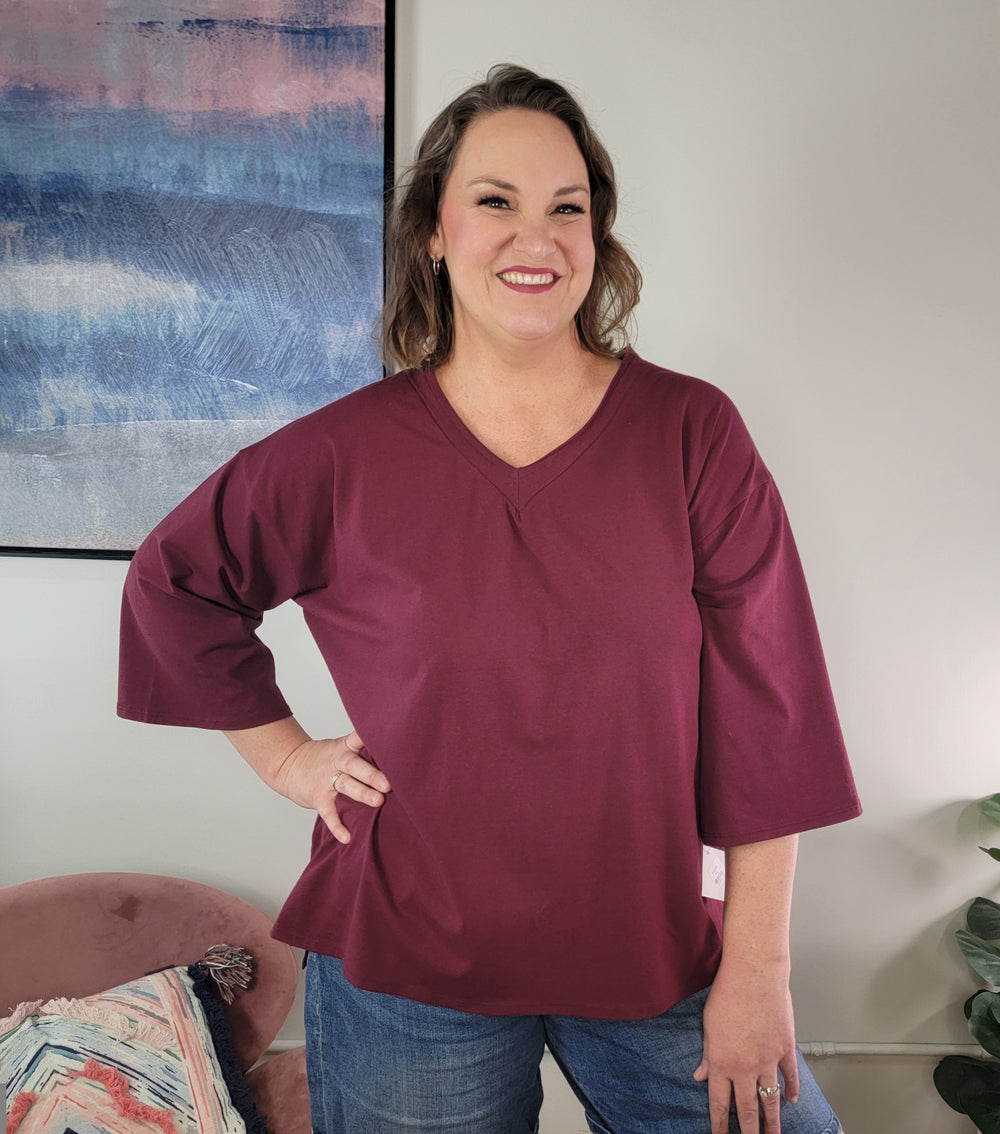A woman with shoulder-length brown hair smiles indoors, wearing the Sew In Love Esme V-neck top in burgundy with wide 3/4 sleeves and blue jeans. A modern abstract painting and a plant are in the background.