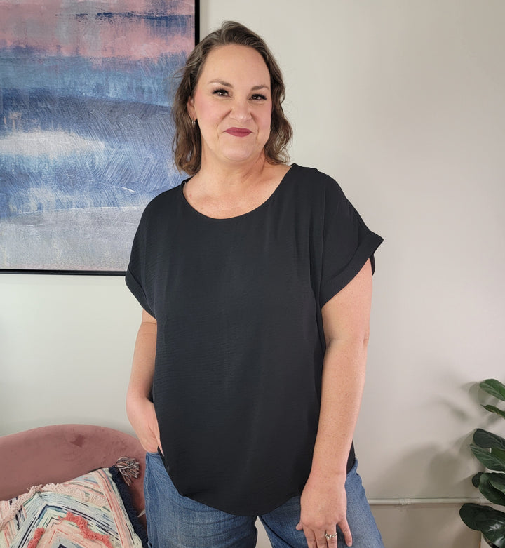 A woman with wavy brown hair smiles in a modern room with abstract art and a plant, wearing blue jeans and the Jamie Blouse from Andree By Unit, with one hand in her pocket.