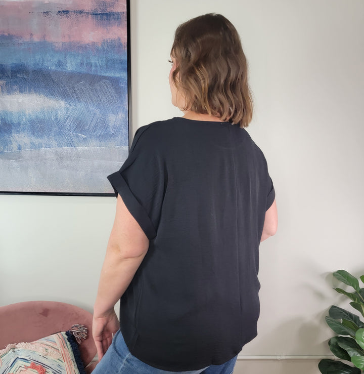 A woman with wavy brown hair stands indoors near an abstract painting and a green plant, wearing the Jamie Blouse by Andree By Unit paired with blue jeans. Her back faces the camera.