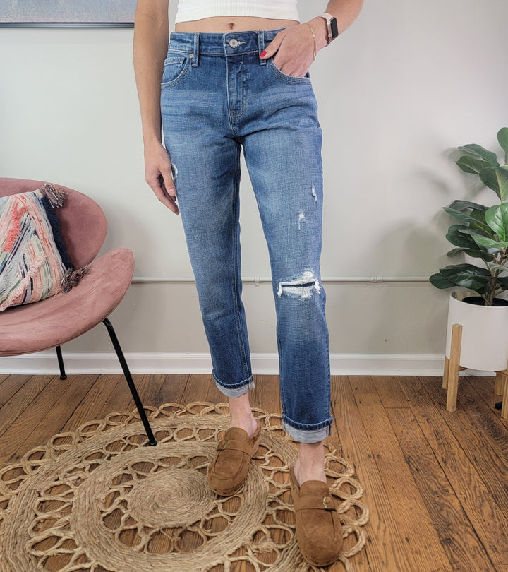 A person stands indoors on a round woven rug wearing KanCan's Lexington Boyfriend Jeans, featuring a subtle rip, paired with brown slip-on shoes. A pink chair and potted plant can be seen in the background.
