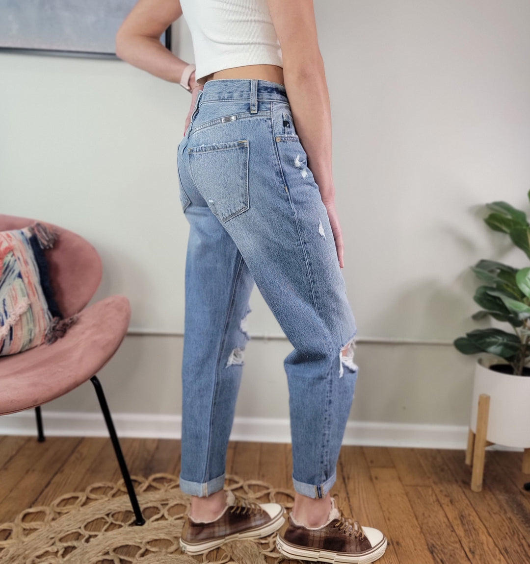 Indoors, a person wears KanCan's Lowell Boyfriend Jeans—light blue, distressed, and cuffed—with a white sleeveless crop top and brown sneakers. A pink chair and potted plant sit on the wooden floor in the background.
