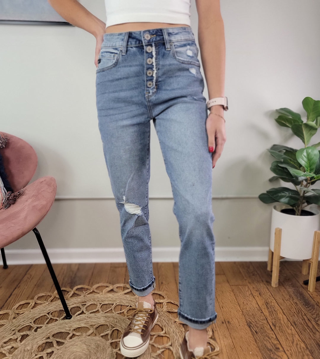 A person wearing the Stormi Mom Jean from KanCan with slight distressing, a white crop top, and brown sneakers stands indoors on a woven rug near a pink chair and a potted plant.
