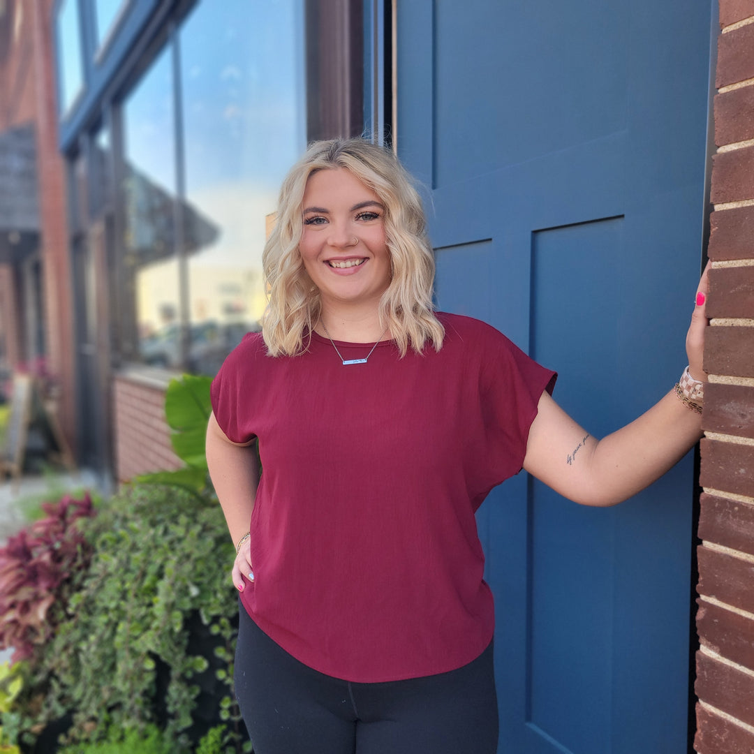 A smiling blonde woman wears the ee:some Brighton Top, a lightweight merlot round-neck style, with black pants as she stands by a blue door and brick wall. Green plants and a window appear in the background.