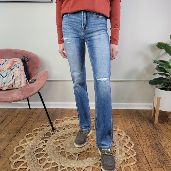 A person wearing a rust long-sleeve shirt, brown loafers, and Risen's Ember Straight Jeans stands on a round woven rug in a room with wooden floors, a pink chair, and a potted plant.
