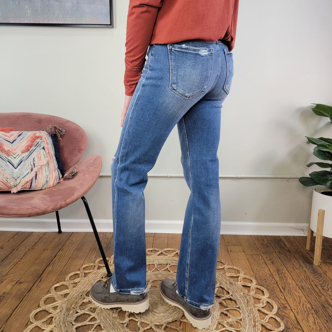 A person in Risen's Ember Straight Jeans, a rust long-sleeve shirt, and gray shoes stands on a circular woven rug near a pink chair with a patterned pillow and a potted plant on a wooden floor.