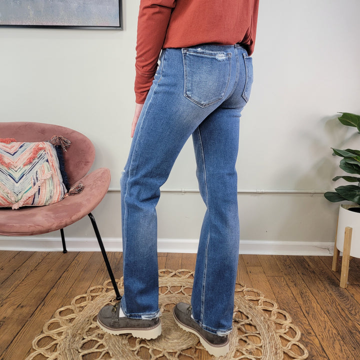 A person in Risen's Ember Straight Jeans, a rust long-sleeve shirt, and gray shoes stands on a circular woven rug near a pink chair with a patterned pillow and a potted plant on a wooden floor.