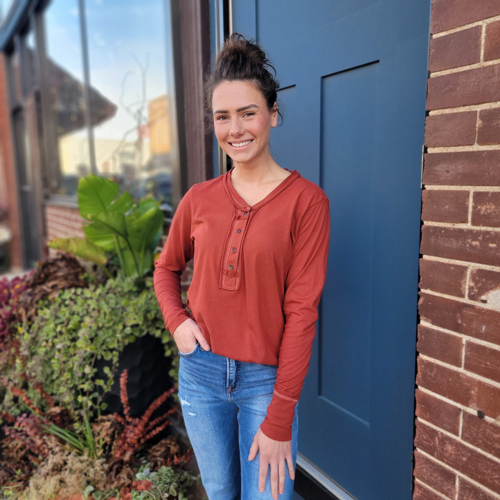 A woman with dark hair in a bun, wearing the ZENANA Della Long Sleeve Top in rust and blue jeans, smiles outside by a blue door and brick wall with nearby plants.