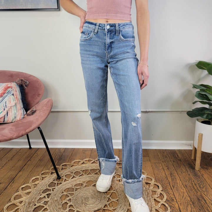 A person wearing Lovervet’s Evermore Straight Jeans in comfort stretch denim with raw hems and white sneakers stands on a circular woven rug, sporting a pink ribbed crop top. A pink chair and plant are visible in the background.