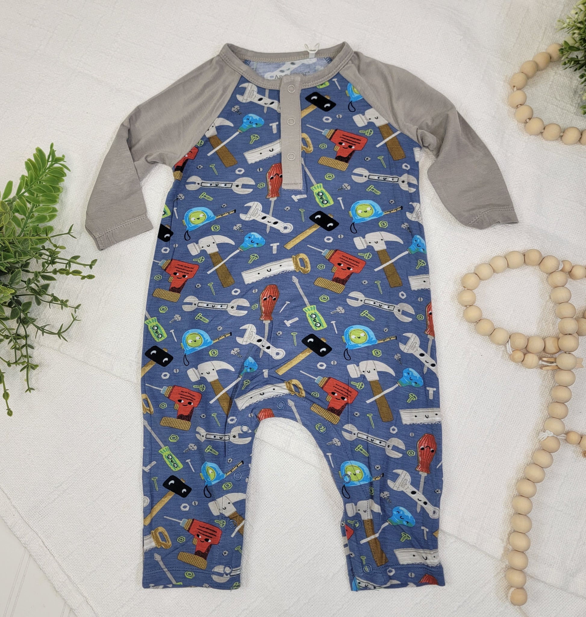 The Angel Dear Kids: Happy Toolbox Romper features gray sleeves, a blue body, and a fun tool print. Designed for easy diaper changes, it's displayed flat with green foliage and wooden beads on a white textured background.
