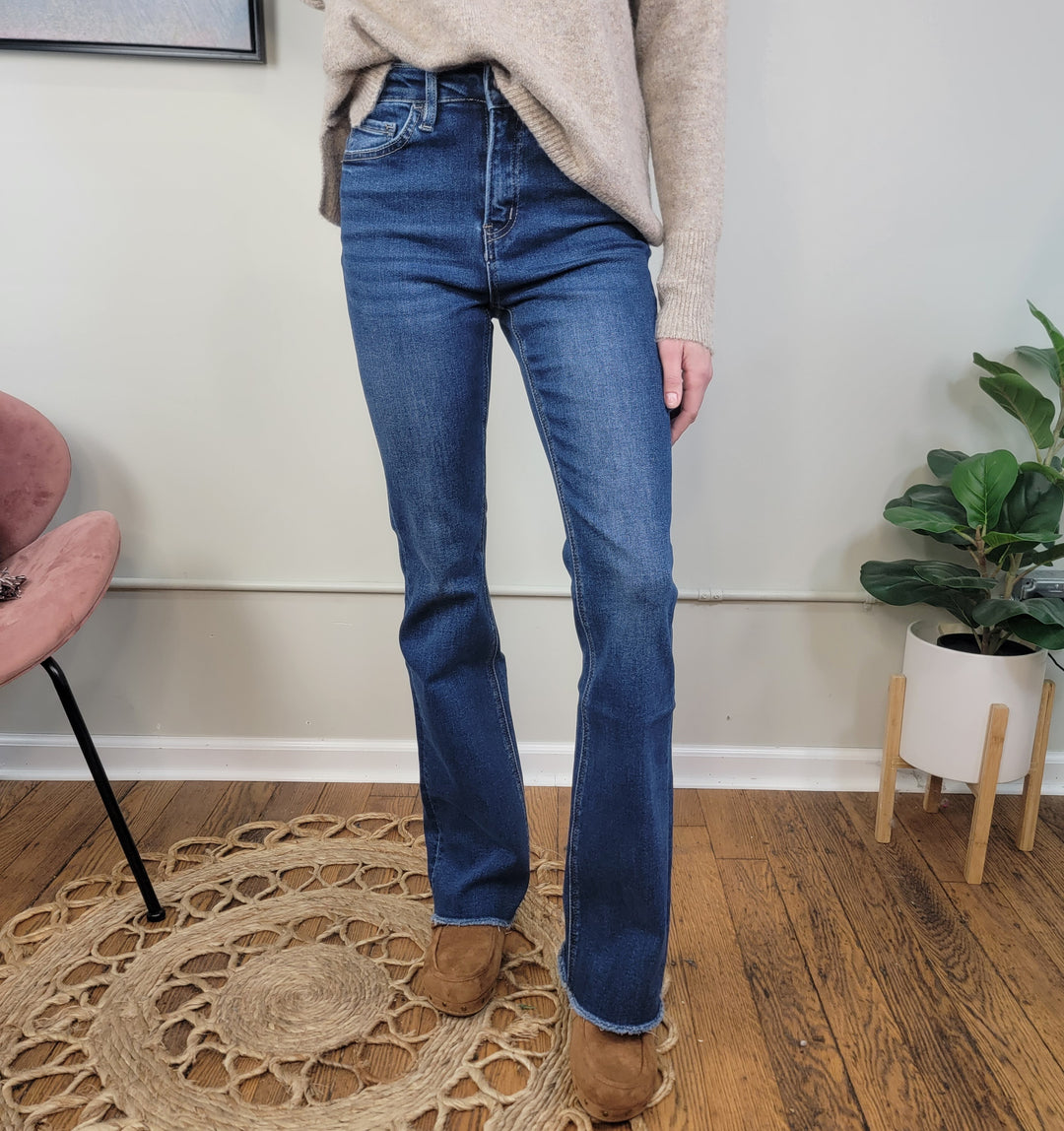 A person wears Lovervet’s Sydney Bootcut Jeans, a beige sweater, and brown shoes while standing on a circular woven rug in a room with wooden floors, a pink chair, and a potted plant. Their head and upper body are out of frame.