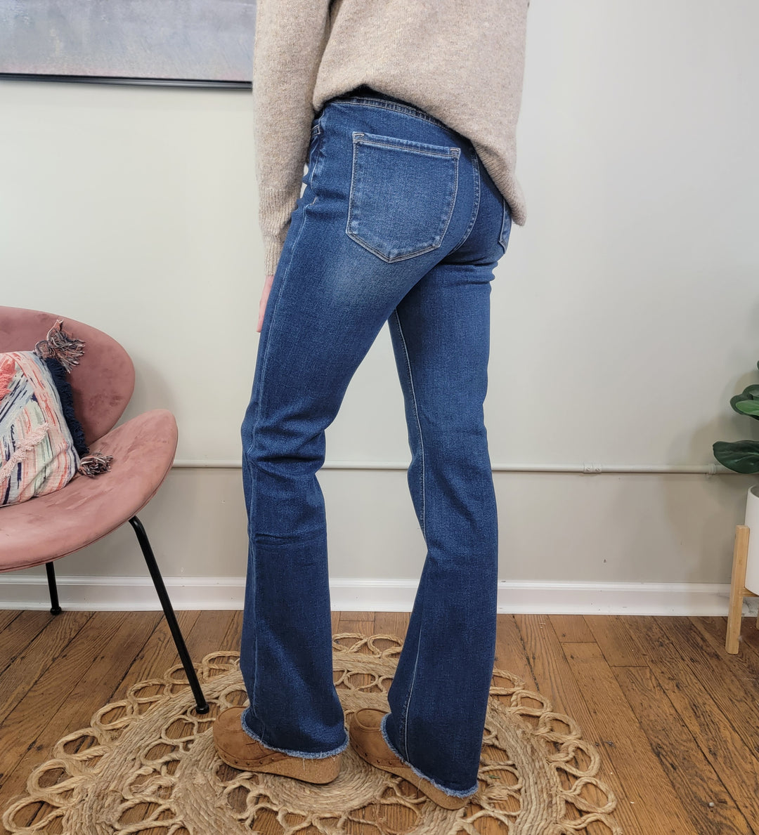 A person stands indoors on a circular woven rug wearing Lovervet's Sydney Bootcut Jeans in blue, paired with a beige sweater and tan shoes. Only their lower half is visible, with a pink chair and plant in the background.