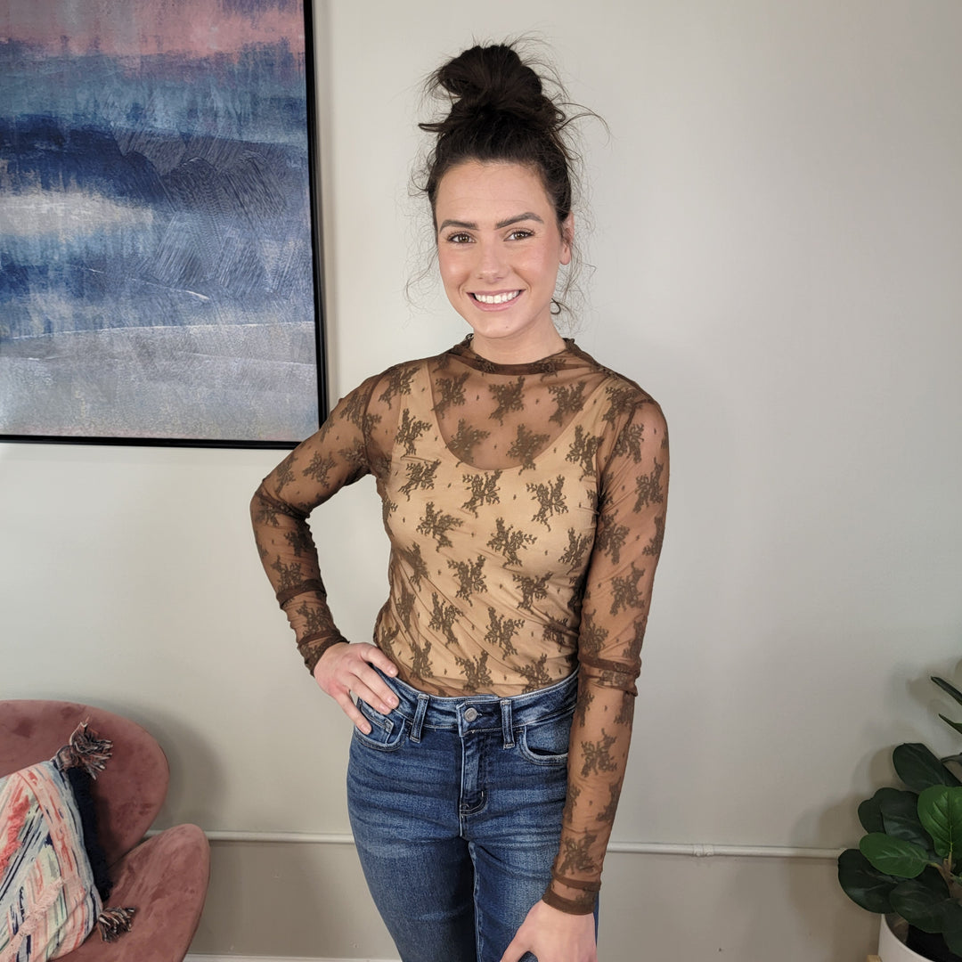 A woman with dark hair in a messy bun smiles indoors, wearing the ee:some CeeCee Brown Lace Top and blue jeans. Art, a pink chair, and a plant add charm to the modern room.