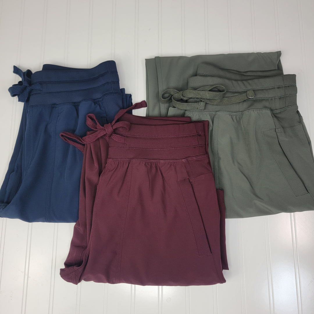 Three pairs of Rae Mode Dance Studio Joggers in navy blue, burgundy, and olive green are folded and stacked on a white textured surface.