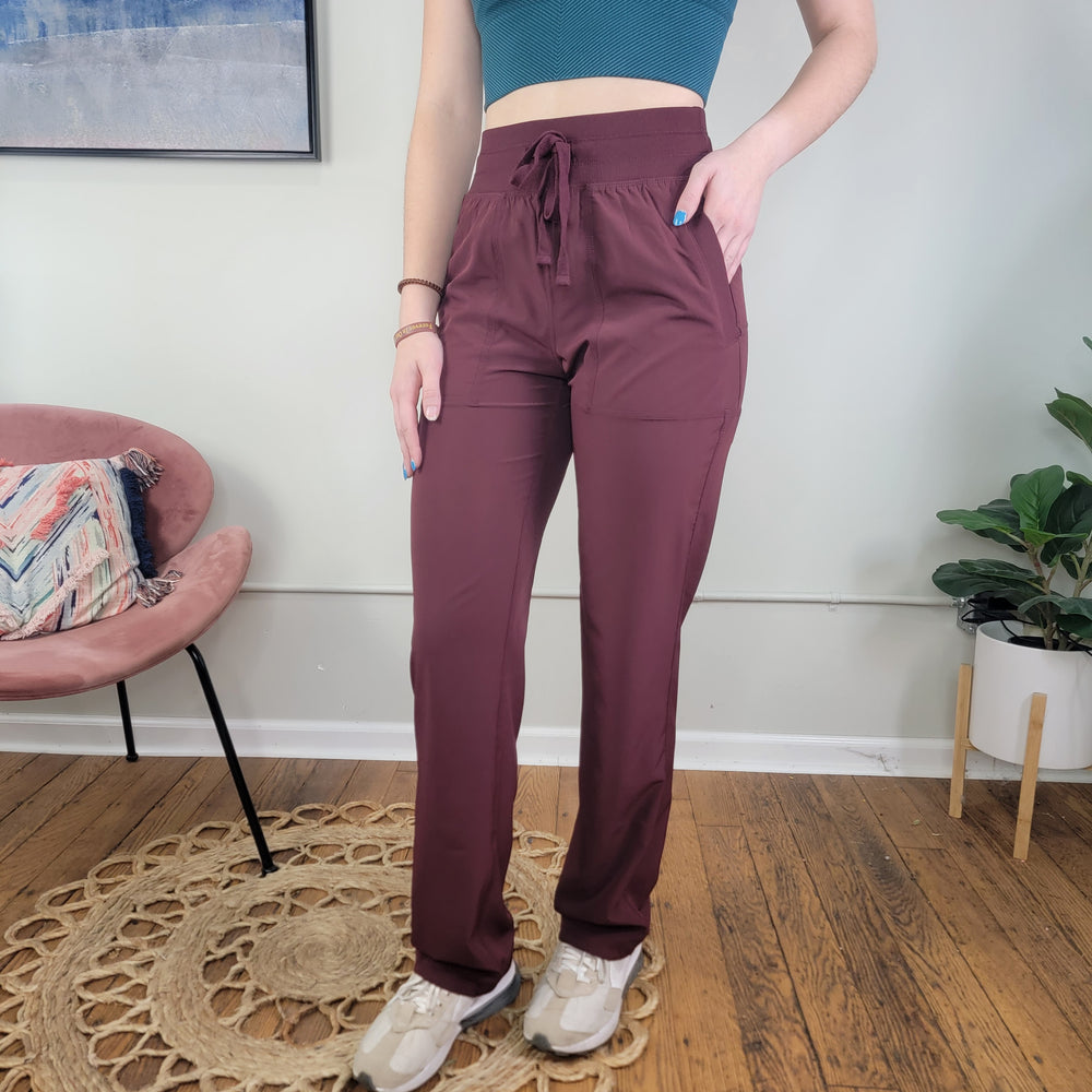 A person in Rae Mode Dance Studio Joggers in maroon, a teal crop top, and white sneakers stands indoors on a wooden floor by a pink chair, potted plant, and wall art. Their face is not visible.