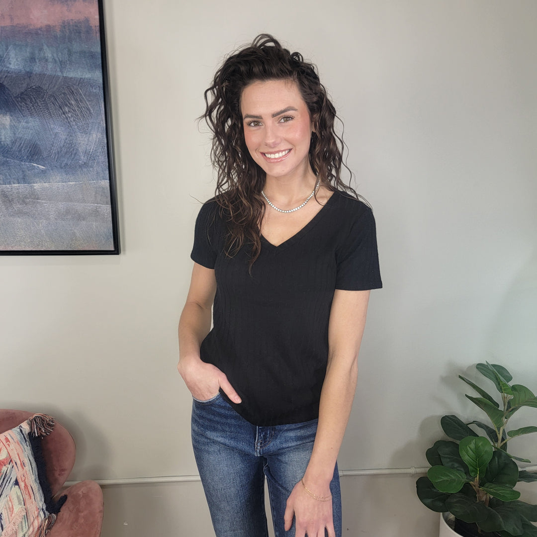 A woman with wavy brown hair is smiling indoors, wearing the ZENANA Nesta V-Neck Top, blue jeans, and a pearl necklace. A plant and a framed picture are in the background.