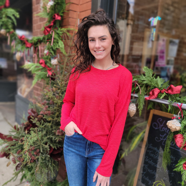 A woman with wavy brown hair, wearing the ZENANA Catalina Long Sleeve Top in bright red and blue jeans, smiles in front of a holiday-decorated storefront with greenery and a chalkboard sign.