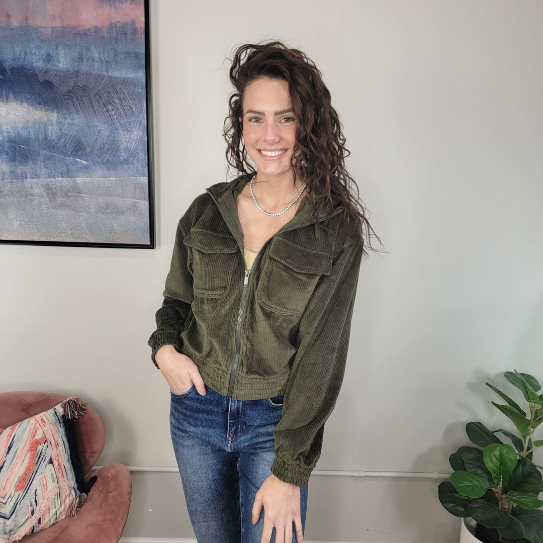 A woman with wavy brown hair smiles indoors wearing the Rae Mode Marilyn Corduroy Jacket in olive and blue jeans. Beside her are a pink chair with a patterned pillow, a leafy plant, and abstract art hanging on the wall.