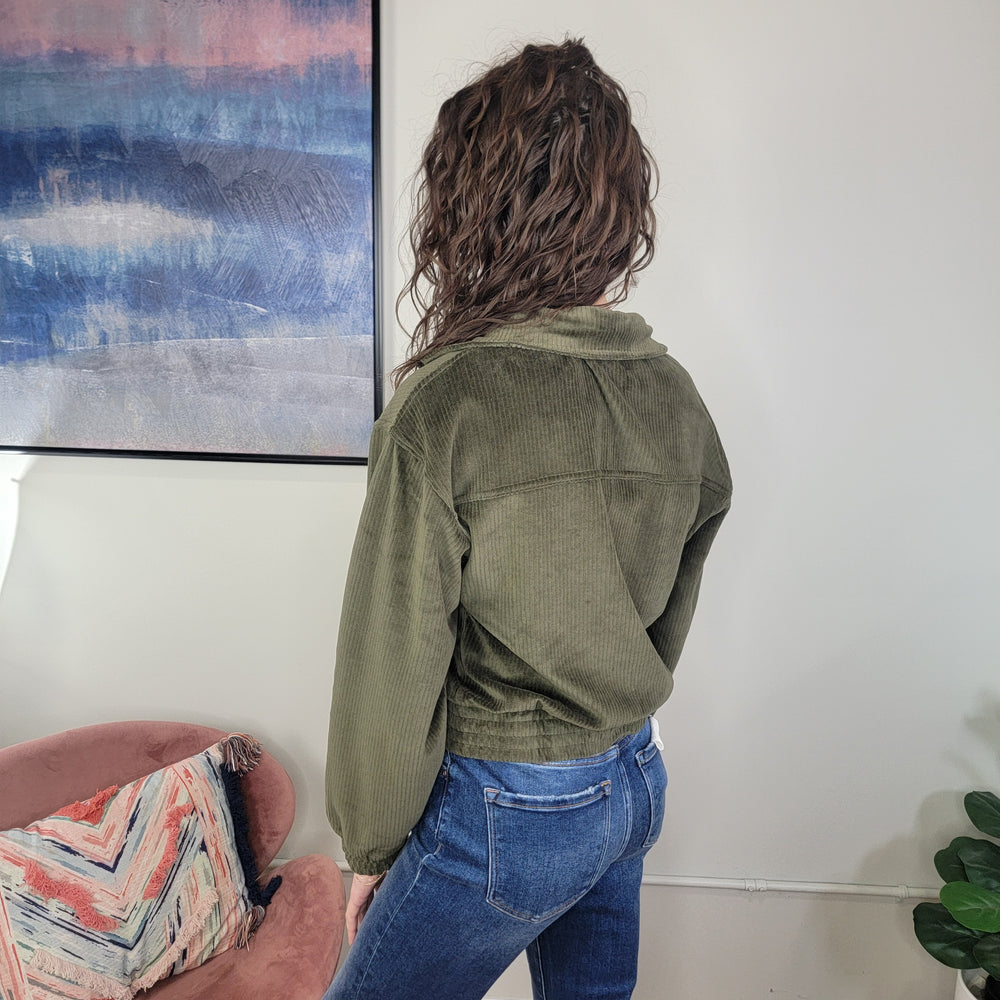 A woman with wavy brown hair stands indoors with her back to the camera, wearing the Rae Mode Marilyn Corduroy Jacket in olive and blue jeans, near a pink chair with a colorful pillow and a large abstract painting.