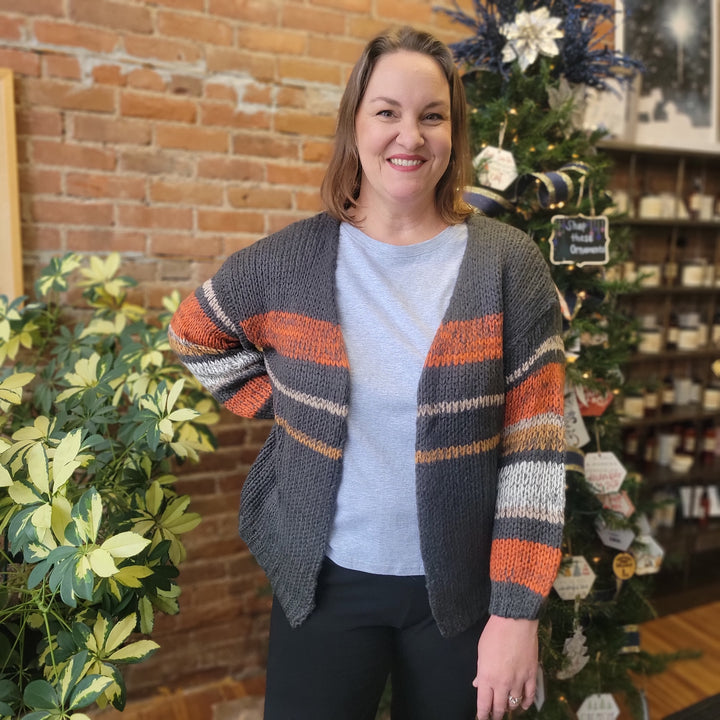 A smiling woman wears the Abigail Stripe Cardigan by Davi &amp; Dani—an orange and beige striped knit with balloon sleeves—standing indoors by a leafy plant and decorated Christmas tree against a brick wall backdrop.