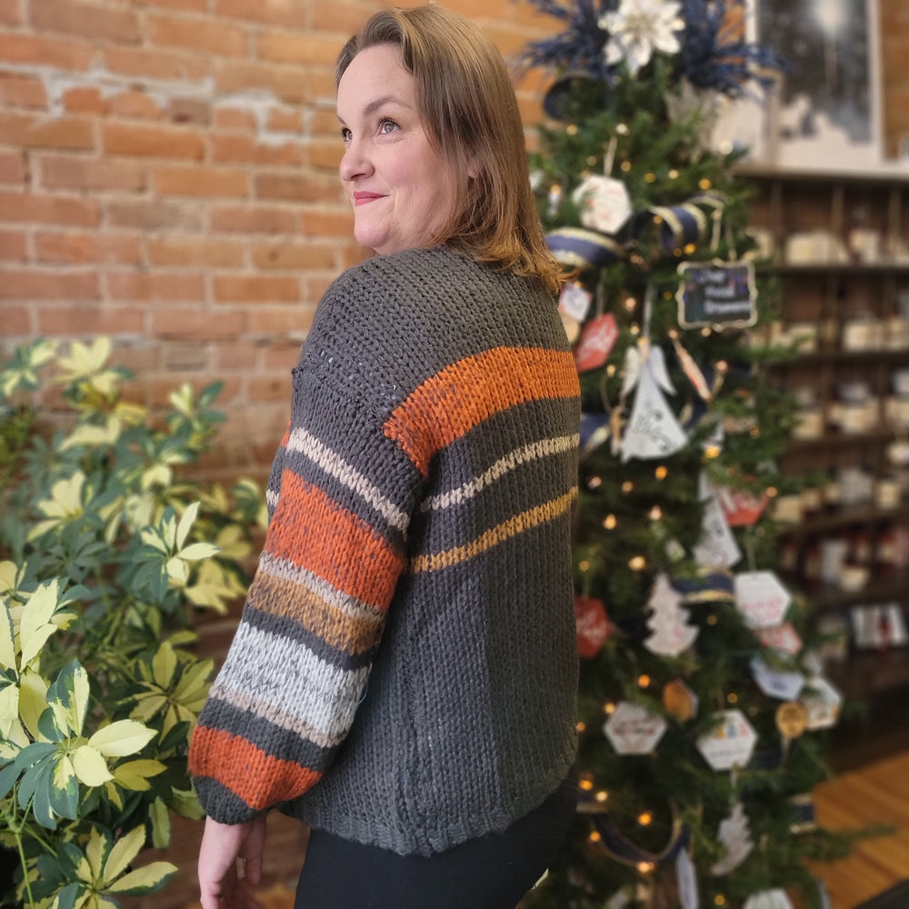 A woman with light brown hair models the Abigail Stripe Cardigan by Davi & Dani, featuring orange, gray, white, and brown striped balloon sleeves, as she stands indoors by a Christmas tree and green plants against a brick wall.
