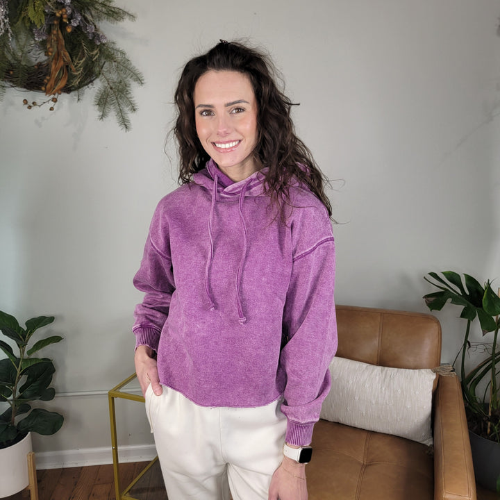 A woman with long brown hair stands indoors, smiling, wearing the ZENANA Lucy Acid Wash Hoodie in purple and white pants. She is next to a tan chair, with green plants and a decorative wreath in the background.