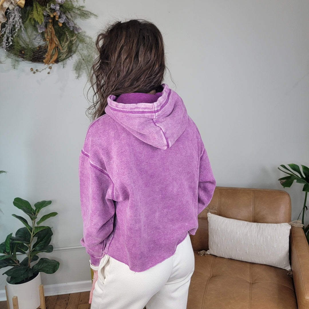 A person with long, wavy brown hair is indoors facing away, wearing the ZENANA Lucy Acid Wash Hoodie in purple and cream pants. Green plants and a tan chair with a pillow are in the background.