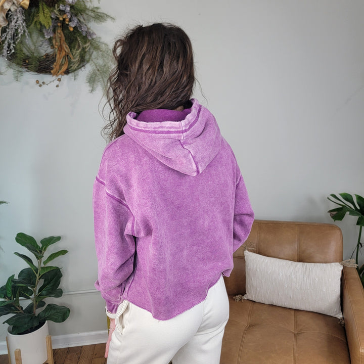 A person with long, wavy brown hair is indoors facing away, wearing the ZENANA Lucy Acid Wash Hoodie in purple and cream pants. Green plants and a tan chair with a pillow are in the background.