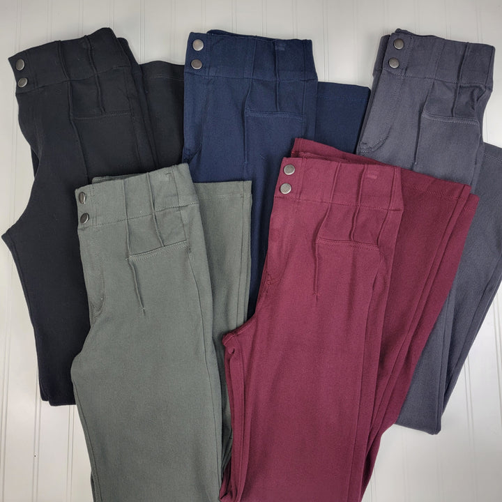 Six pairs of Rae Mode Landry Stretch Dress Pants, each high-waisted with two metal buttons, are neatly arranged side by side in black, dark green, navy blue, burgundy, charcoal gray, and light gray on a white surface.