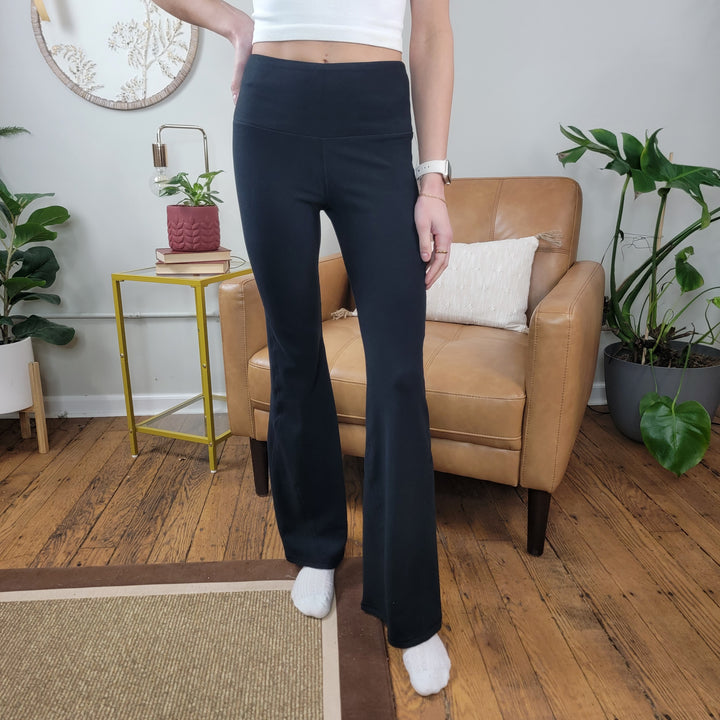 A woman stands indoors on a wooden floor, wearing Mono B's Malison Flared Fleece Leggings paired with a white crop top and white socks. She is in front of a tan armchair, with green plants and a small yellow table nearby.