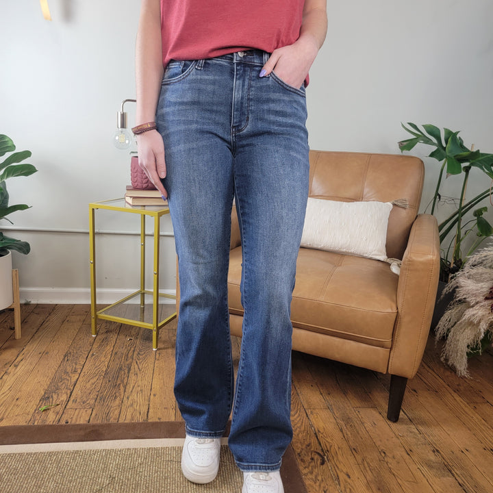 Someone wearing Crosby Thermal Judy Blue Bootcut Jeans by Judy Blue, white sneakers, and a red top stands on a wooden floor with one hand in their pocket. Behind them are a tan armchair, green plants, and a yellow side table with a lamp.