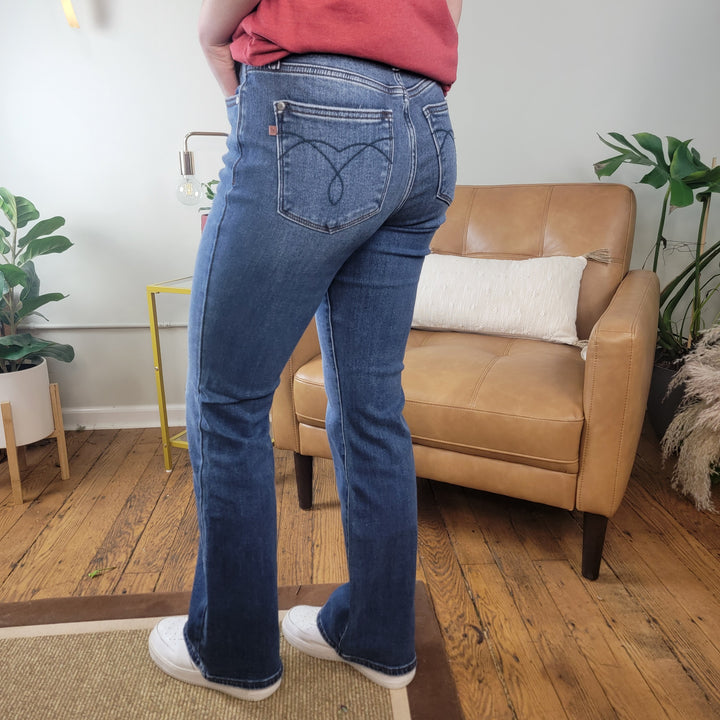 A person stands indoors on a wooden floor near a tan leather chair, wearing Crosby Thermal Judy Blue Bootcut Jeans by Judy Blue, a red shirt, and white sneakers. Green plants and a yellow side table are visible in the background. Photo taken from behind.