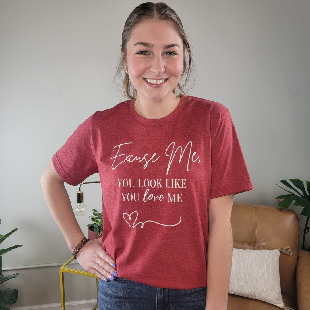 A smiling woman stands indoors wearing the Cosmos + Clover Boutique "You Look Like You Love Me" Graphic Tee in heather red, featuring a heart design and text. She poses with one hand on her hip in front of a tan sofa and small table.