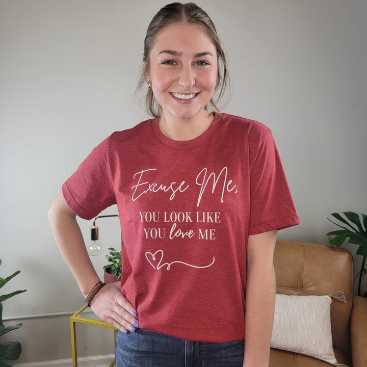 A smiling woman stands indoors wearing the Cosmos + Clover Boutique "You Look Like You Love Me" Graphic Tee in heather red, featuring a heart design and text. She poses with one hand on her hip in front of a tan sofa and small table.