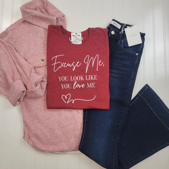 A flat lay on a white background features a pink plaid button-up shirt, the Cosmos + Clover Boutique "You Look Like You Love Me Graphic Tee" in heather red, and dark blue jeans with tags.