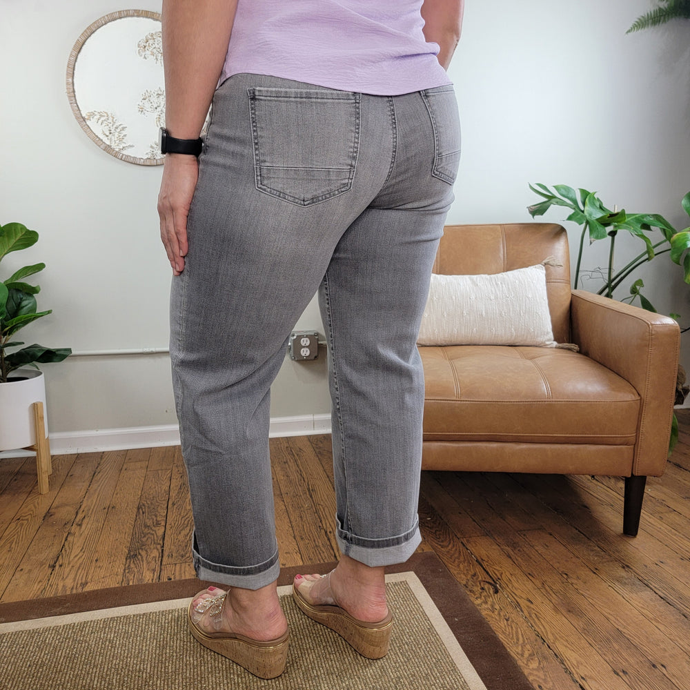 A person wearing Kut from the Kloth's Charlotte Explained Jean and a light purple top stands with their back to the camera in a cozy, plant-filled living room with a tan chair and wood floors.