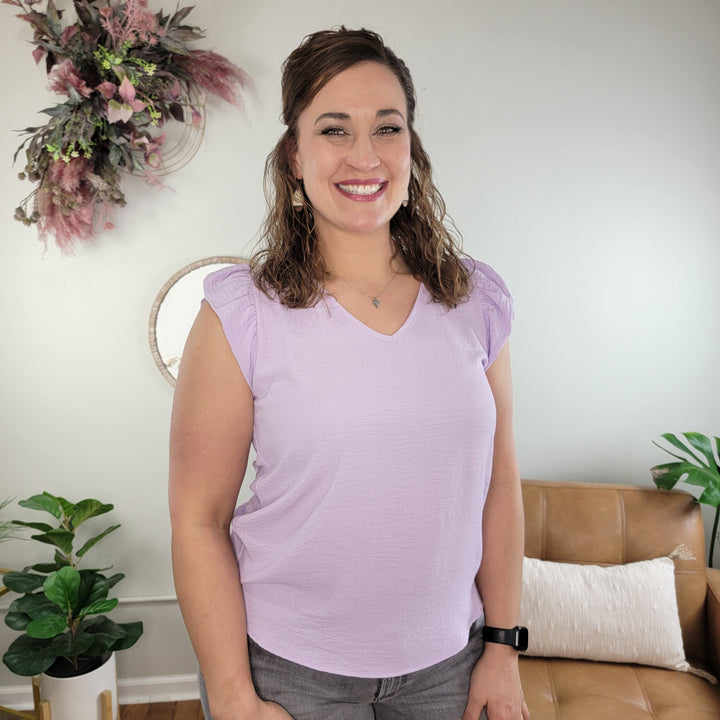 A woman with wavy brown hair smiles in a bright living room, wearing gray pants and the Les Amis Presley V-neck Top—a lavender ruffle sleeve blouse made of lightweight polyester. Plants and a decorative wreath adorn the space.