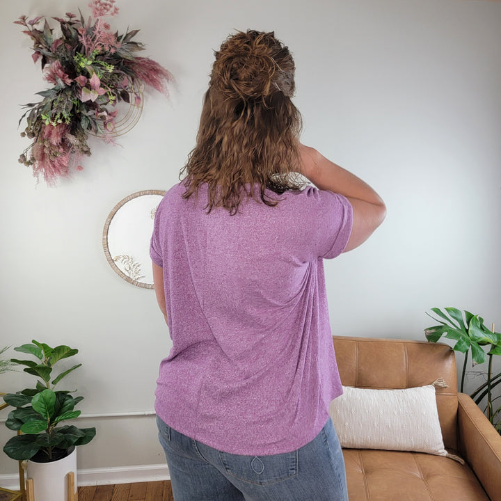 A person with wavy, shoulder-length hair wears the ZENANA Tania Top in a cozy living room decorated with plants, a tan couch, dried flowers, and a round mirror.