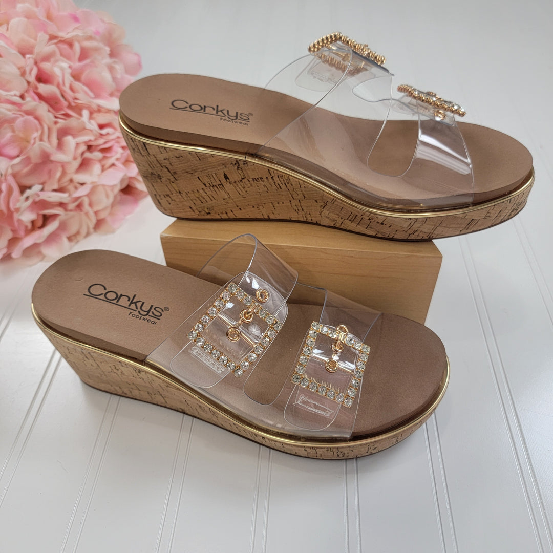 The Corkys Twinsie Wedge features cork soles, clear straps, gold and rhinestone buckle accents, and a memory foam footbed. Displayed on a white surface next to pink flowers.