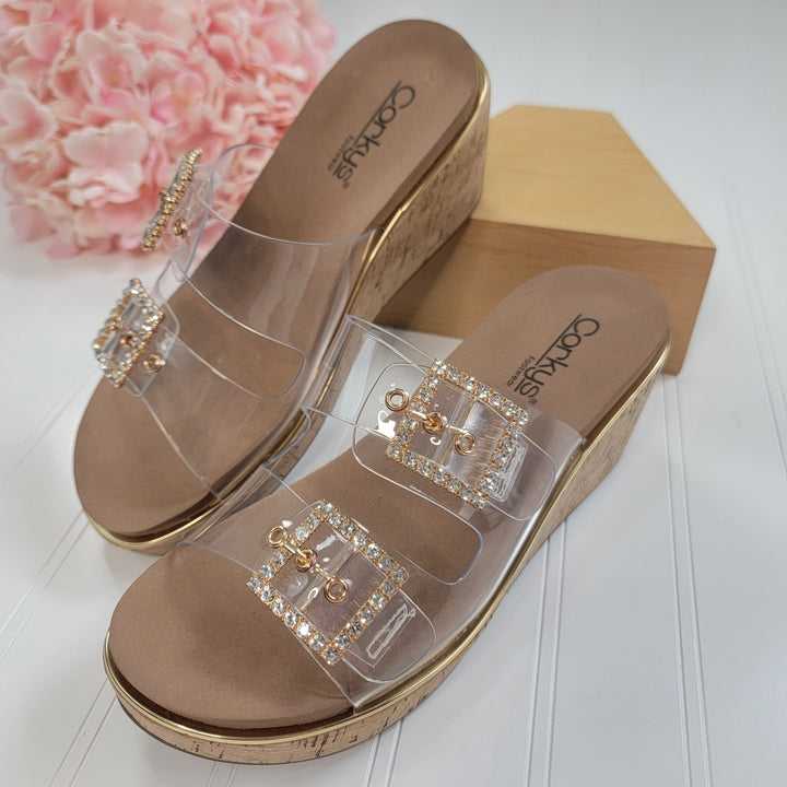 The Twinsie Wedge from Corkys features clear straps, a memory foam footbed, and rhinestone-encrusted buckles. Displayed on a white surface with a wooden block and pink flowers in the background.