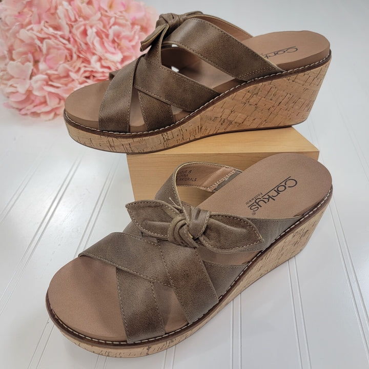 A pair of Corkys Bowdacious Sandals in taupe with criss-cross straps and a bow, featuring a memory foam insole, is displayed on a white surface with a light wooden block and pink flowers in the background.