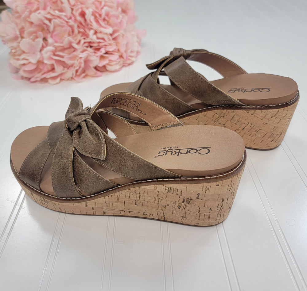 The Bowdacious Sandal from Corkys features tan suede bow-tied straps, cork wedge soles, and a memory foam insole. Displayed on a white surface with pink flowers in the background.