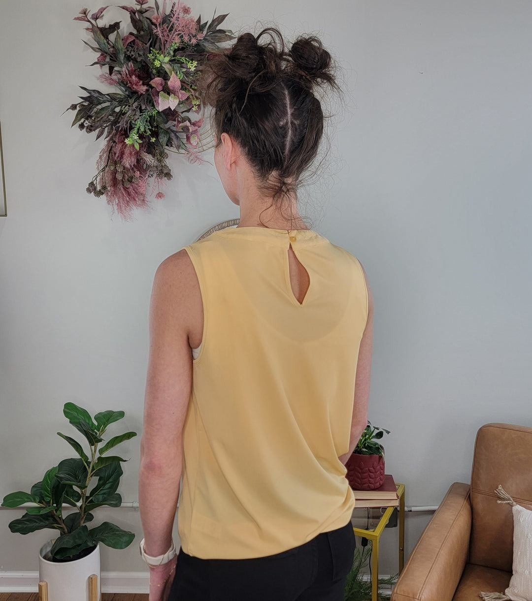 A person with dark hair in two buns models the Doe &amp; Rae Kaelie Pleated Sleeveless Top and black pants, standing with their back to the camera in a living room with plants, a beige couch, and a floral wall arrangement.