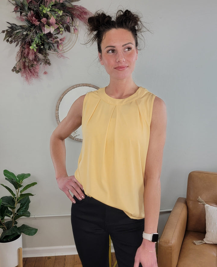 A woman with dark hair in two buns wears the Kaelie Pleated Sleeveless Top by Doe &amp; Rae, a dusty yellow, flattering sleeveless piece, paired with black pants. Indoors near a tan armchair and plants, she smiles slightly while looking to the side.