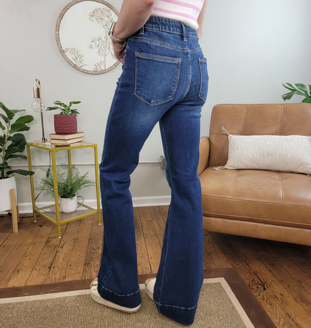 A person wears Risen's Selah Tummy Control Flares - Dark Wash jeans and sneakers, standing on a rug in a cozy living room with a brown sofa, plants, and a small yellow side table stacked with books.